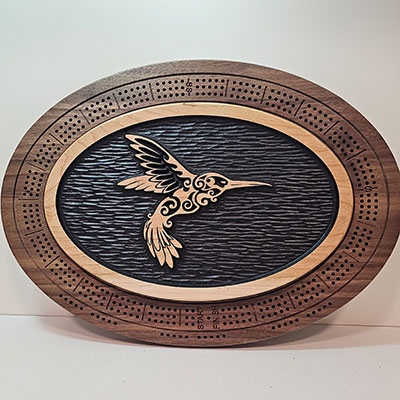 Oval cribbage board with a carved, patterned hummingbird at center, framed by detailed wooden scoring holes.