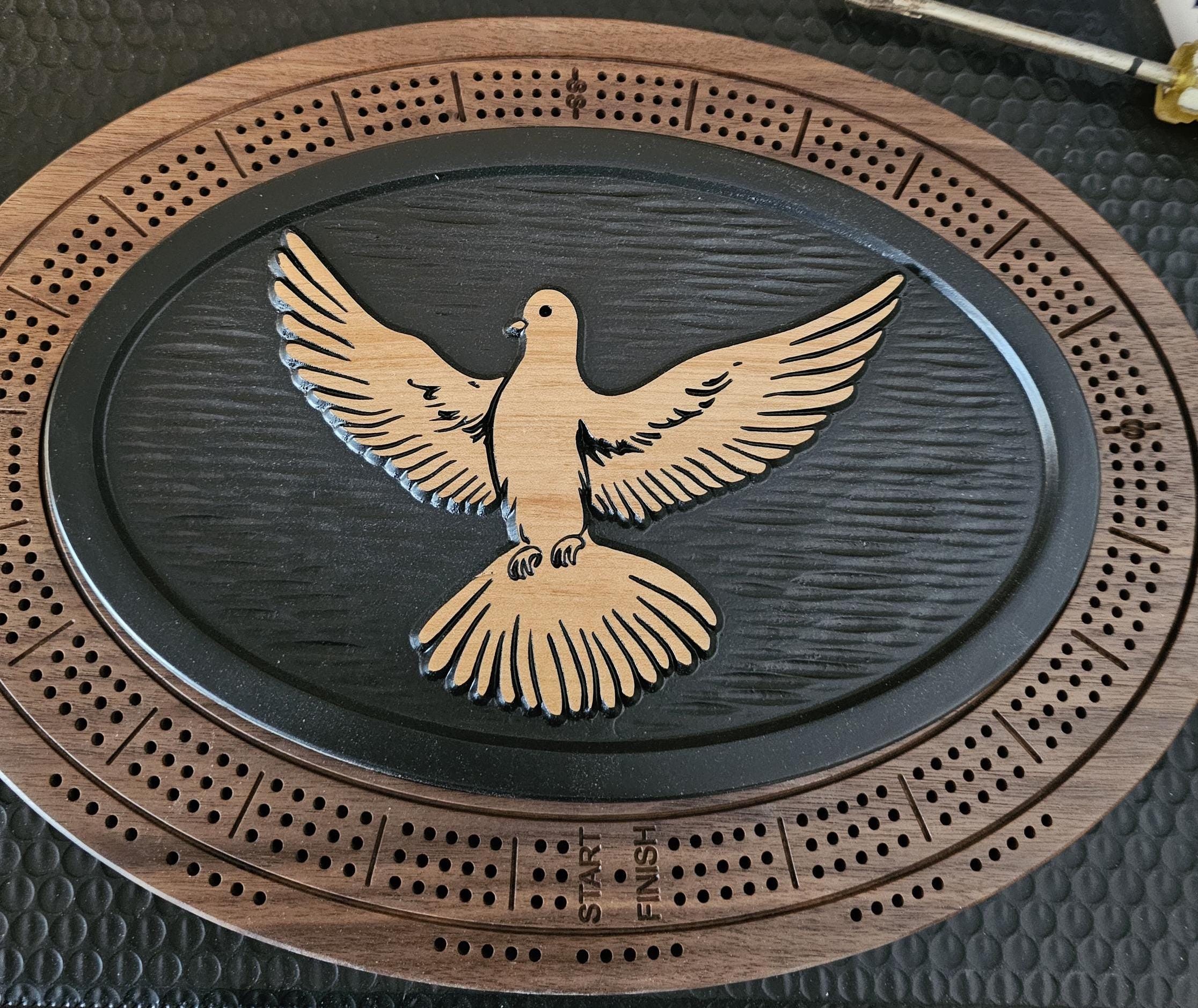 Oval wooden cribbage board with carved dove design on dark stained center and natural wood border